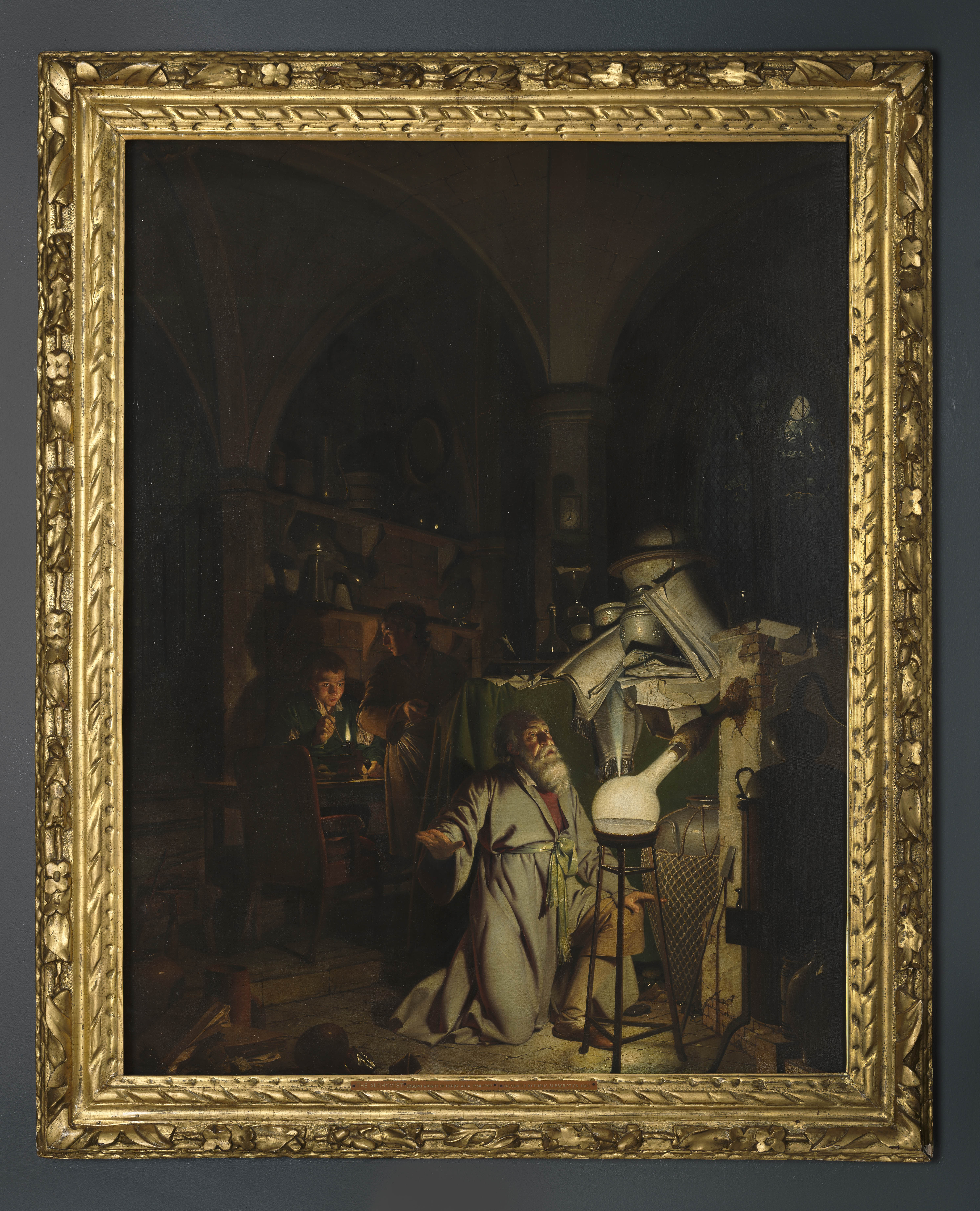 Painting of an alchemist kneeling before a glowing glass flask from which a jet of gas is being released. To his left two young, male assistants are working with a bunsen burner. One of them has stopped to turn and point towards the alchemist. The scene is set within a dark interior, the architecture of which appears gothic. Beyond the alchemist, through an arched, leaded window, a full moon is visible. Inscribed recto, to right of centre: 'J.W. /Pinxt. /1795'. Painted by Joseph Wright of Derby (1734 - 1797). Oil on canvas, in carved and gilded frame. Glazed.
