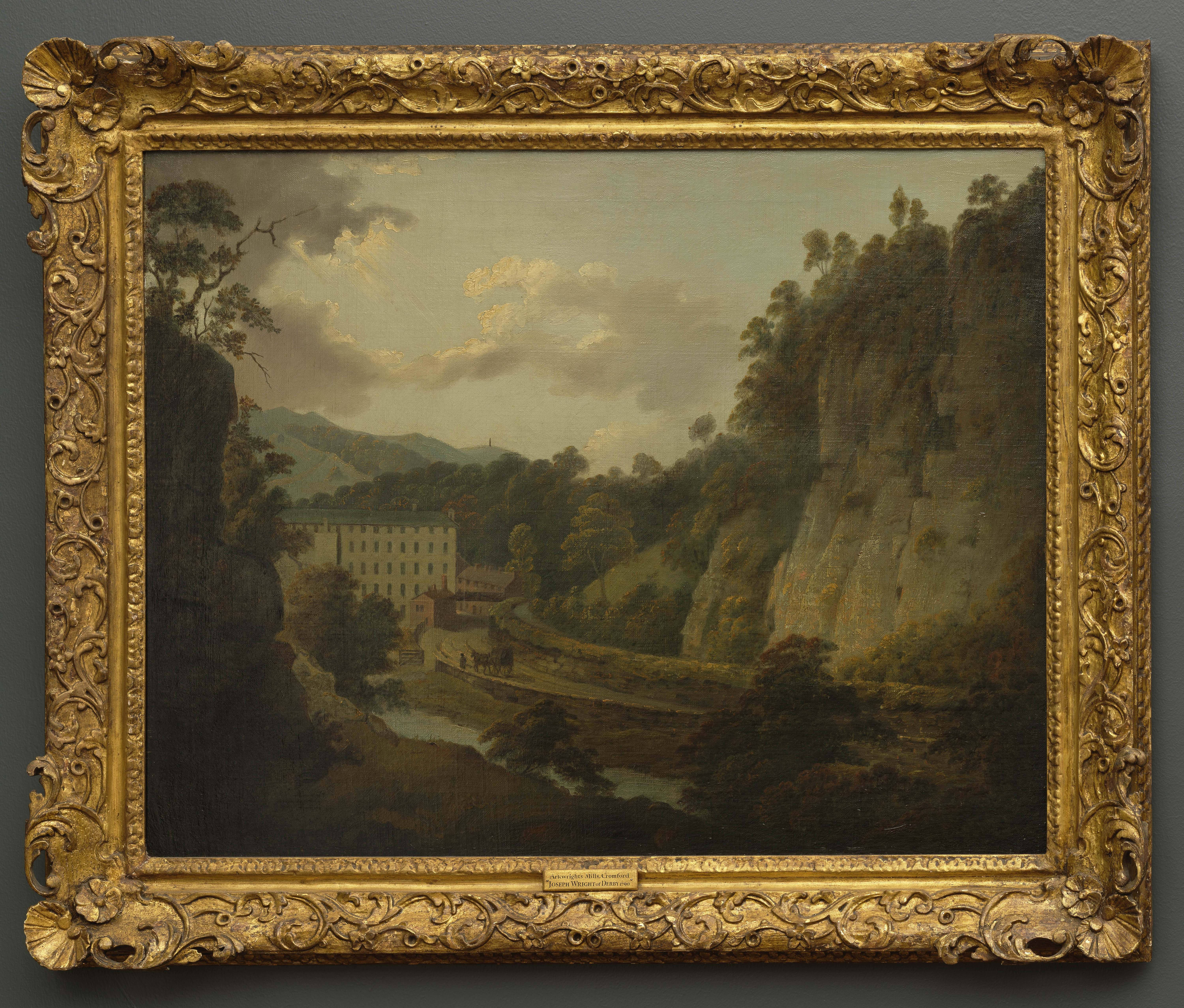 Painting of a landscape identified as Arkwright's Mills at Cromford. The mills appear nestled within a high-sided gorge. A road and brook lead towards them. Beyond, green hills and tall monument can be seen. Sunlight streams through clouds to the left of the scene. Painted by Joseph Wright of Derby (1734 - 1797). Oil on canvas in gilded frame.